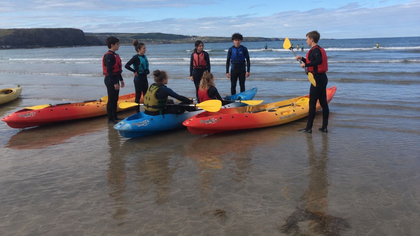 Alumnos con chaleco salvavidas escuchan a su monitor en una playa, junto a unos kayaks