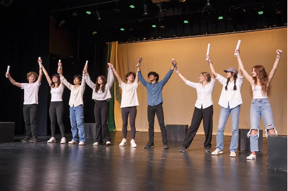 Nueve alumnos con los brazos en alto saludando sobre un escenario